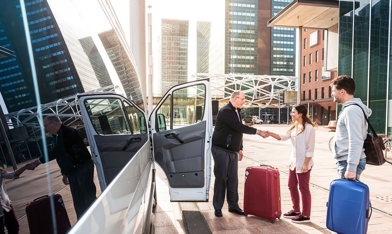 Driver of a airport shuttle minivan, greeting his passengers with their luggage on the sidewalk of a modern city business district